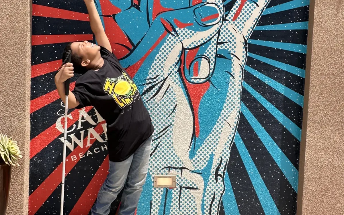 Grayson Roberts is standing with his cane and looking up into the sky. His left arm to stretch up with a "rock on" hand gesture. The background is a colorful "rock on" fingers image.