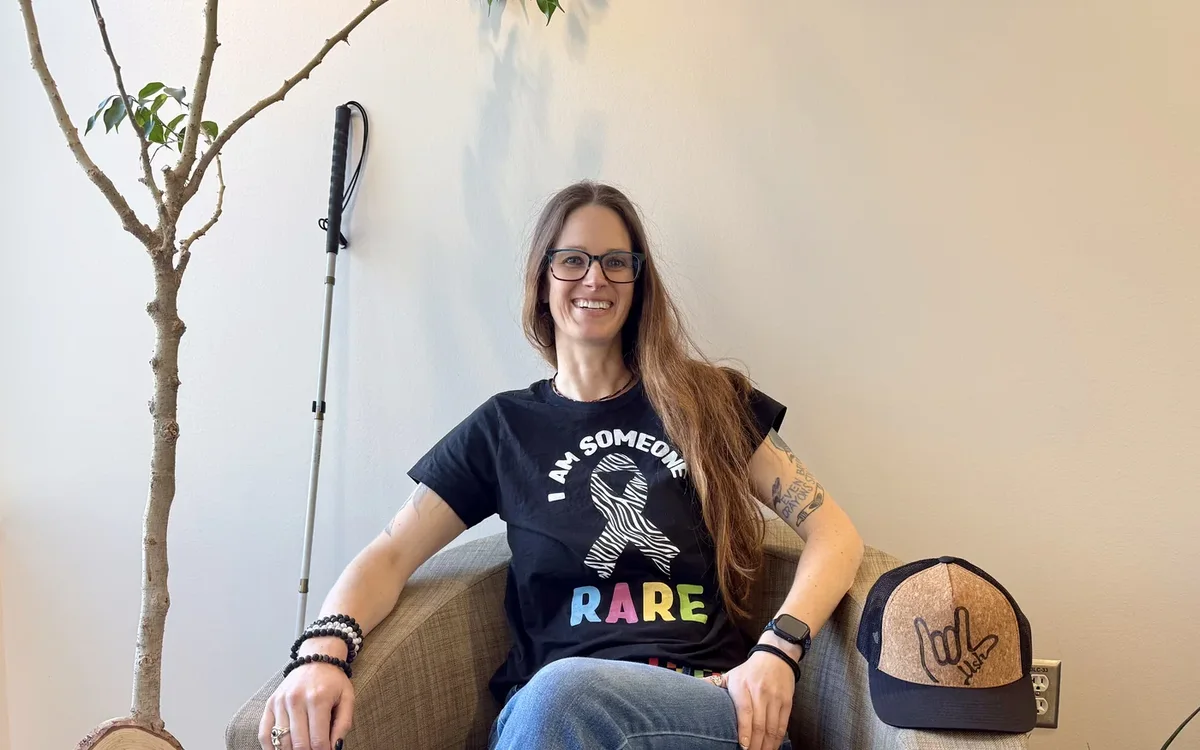 Krista Webb sitting in a woven armchair in a black “I am someone RARE” T-shirt, a white cane leaning on the wall beside her.