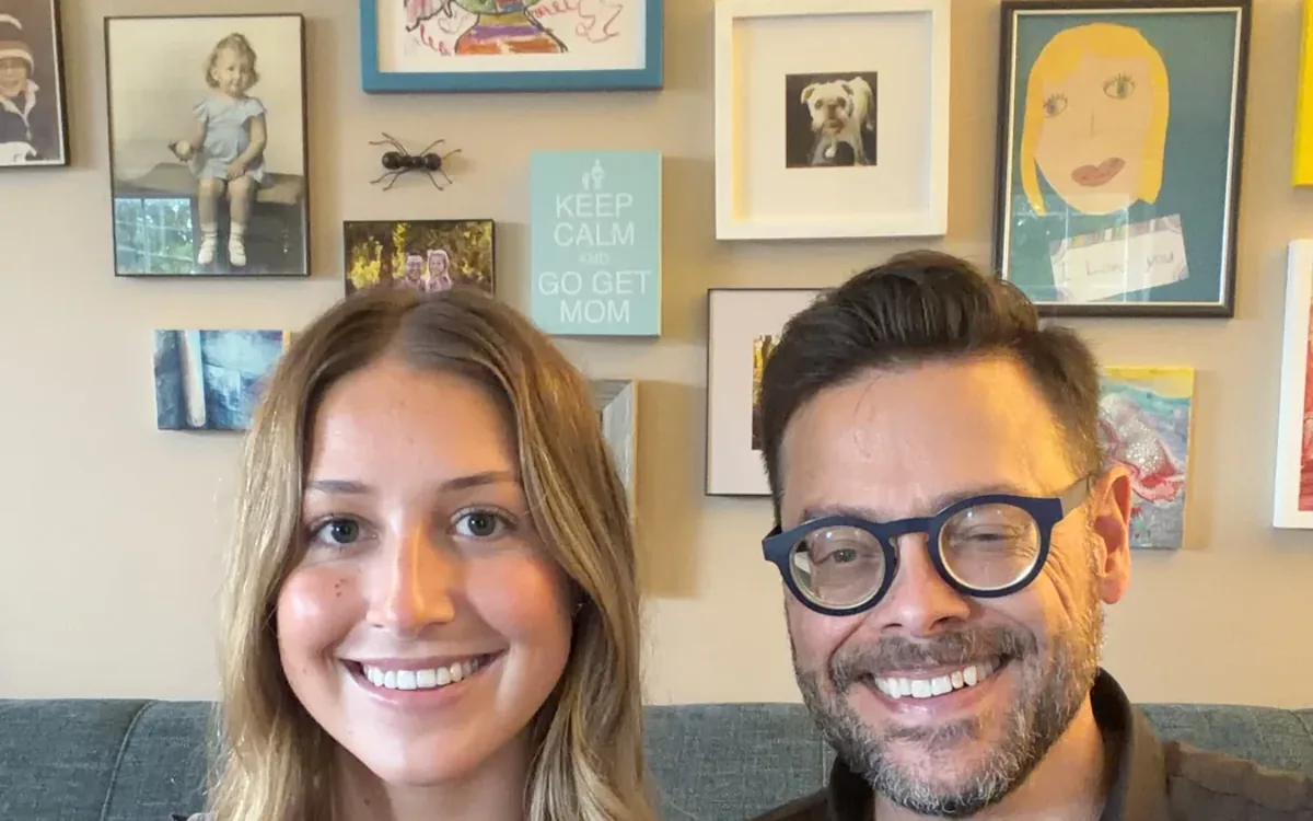Father and daughter, Josh and Eileen Loebner excitedly smiling, sitting together on a couch with whimsical family artwork framed on the wall behind them.