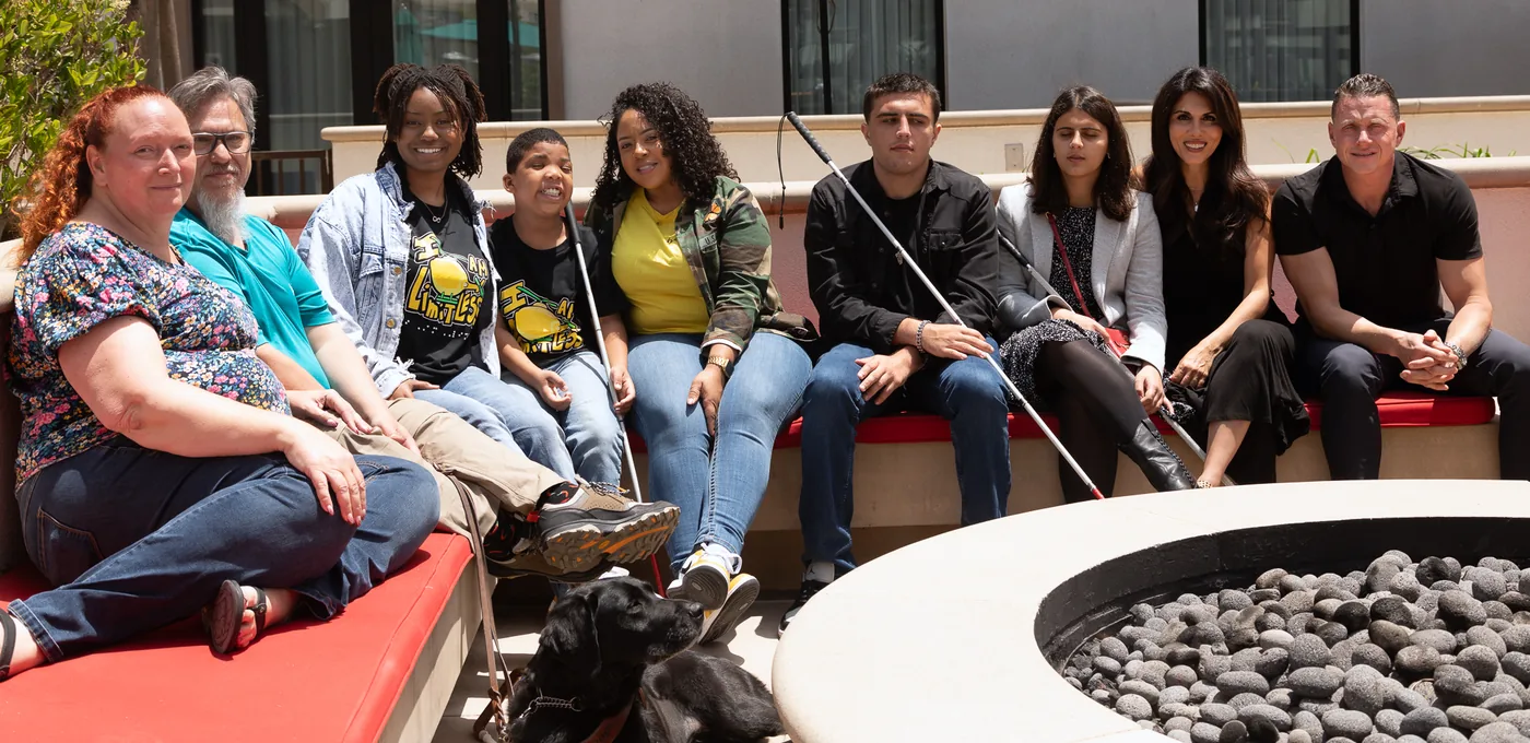 Grant winners and their families sitting together along a low outdoor bench beside a fire pit. Several hold white canes and a black guide dog rests in front of them.