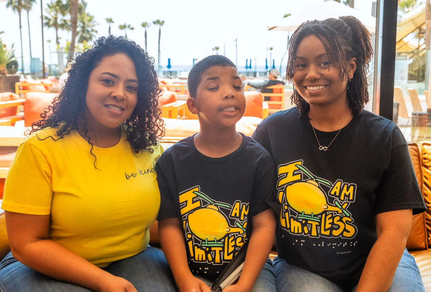 Three members of Team Limitless sitting together on a sofa by a window, two of them in black “I AM LIMITLESS” T-shirts, palm trees outside behind them.