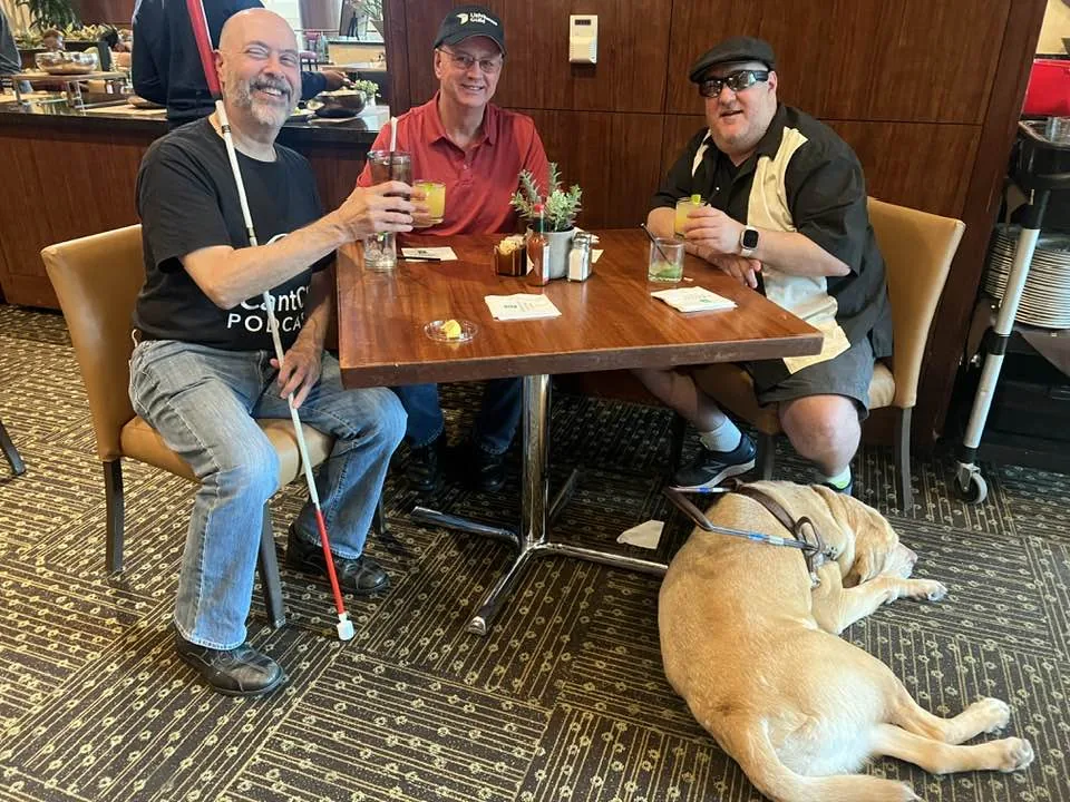 David, Ed, and Brian are doing a cheers at dinner. Wesley, Brian's guide dog is resting under the table.