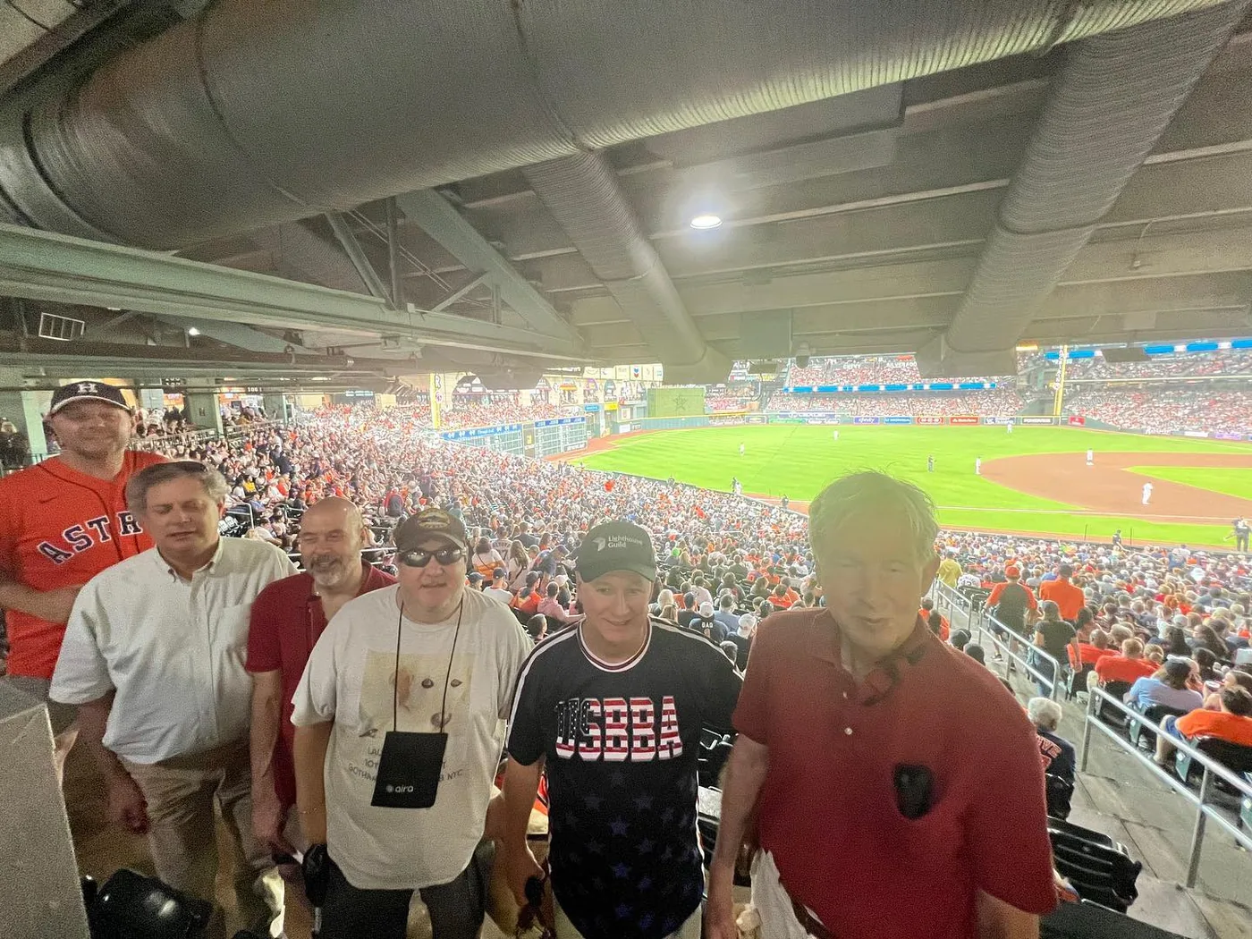 David, Brian, Ed, and Mike are at the Houston Astros game. They are with two other people. The field is in the background.