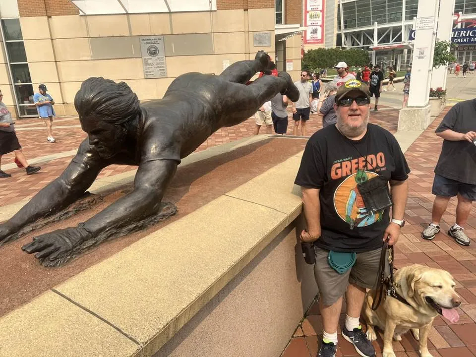 Brian standing with his guide dog Wesley in front of a statue of Pete Rose sliding.