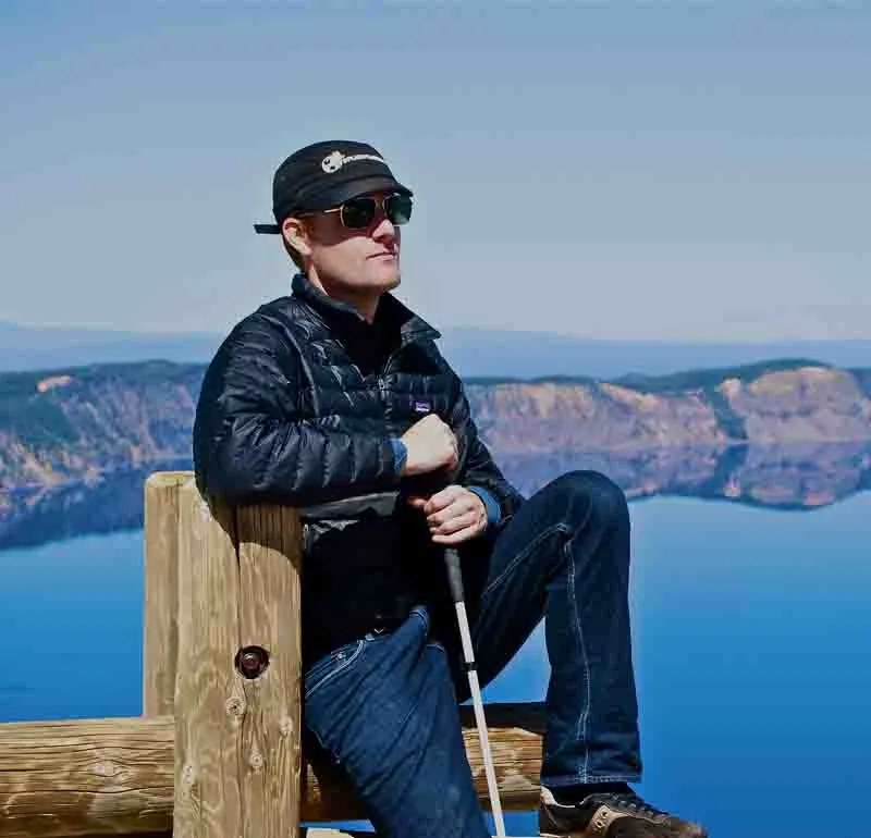 Mike is sitting on a fence at Crater Lake holding his cane.