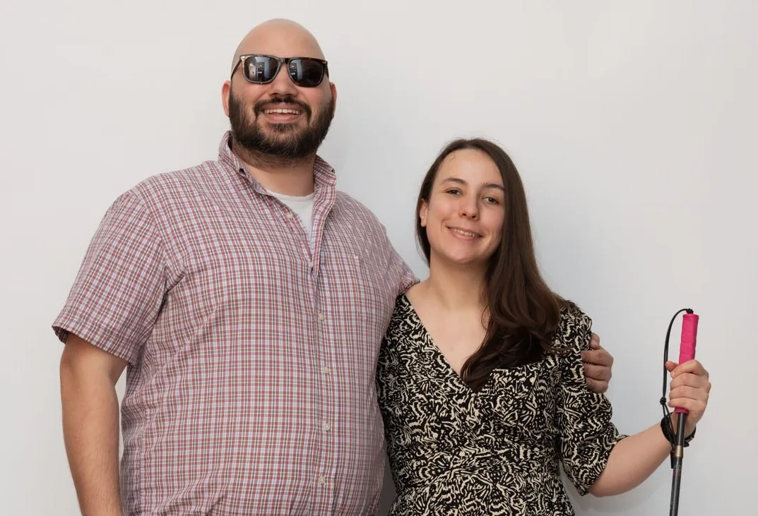 Emeline Lakrout and Daniel Gutz standing arm in arm against a plain pale backdrop. He wears dark glasses and a checked shirt; she holds a white cane with a pink grip.