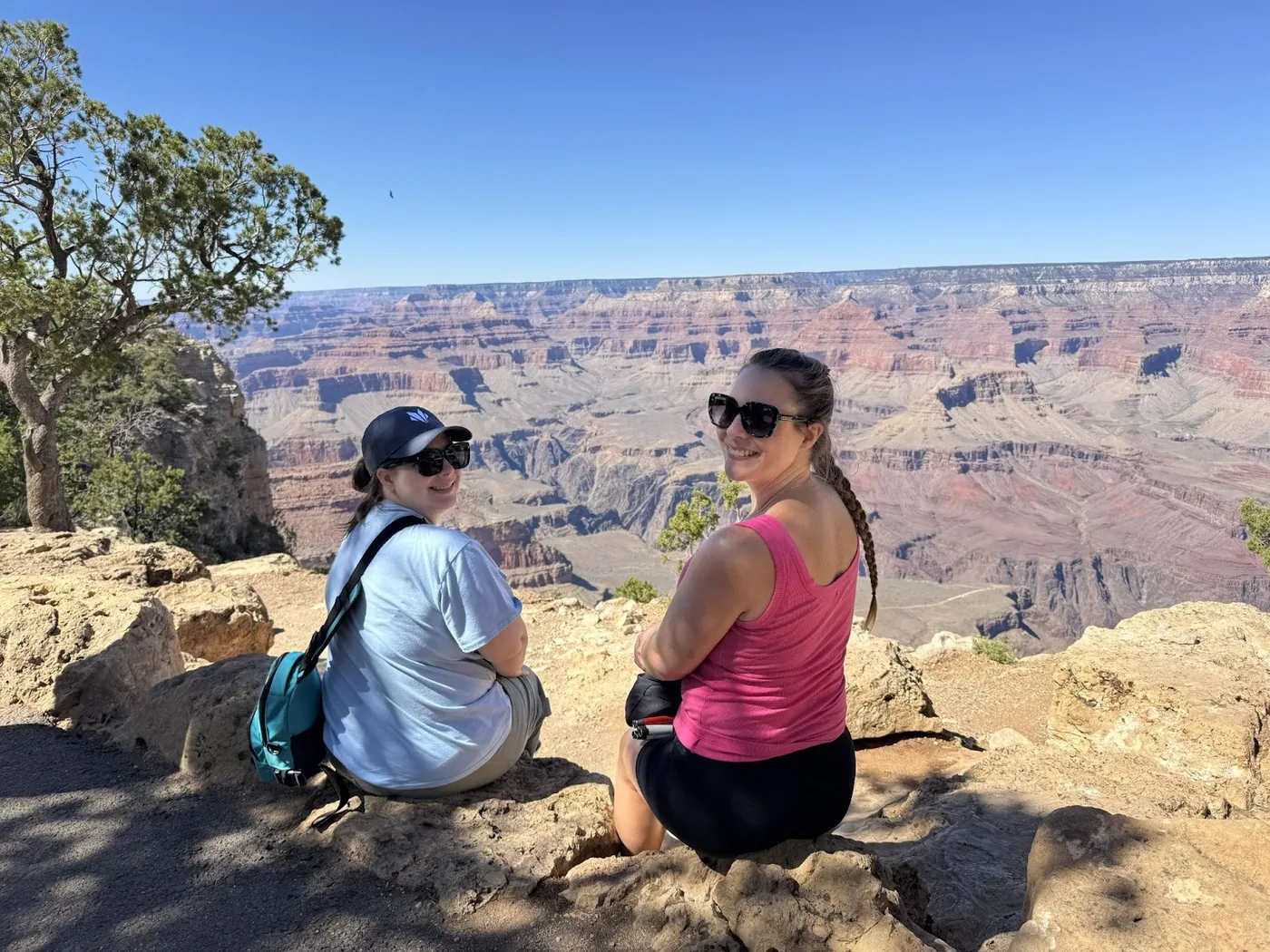 A photo of Erin and Monica sitting on rocks, facing the Grand Canyon, but turning back to smile towards the camera.