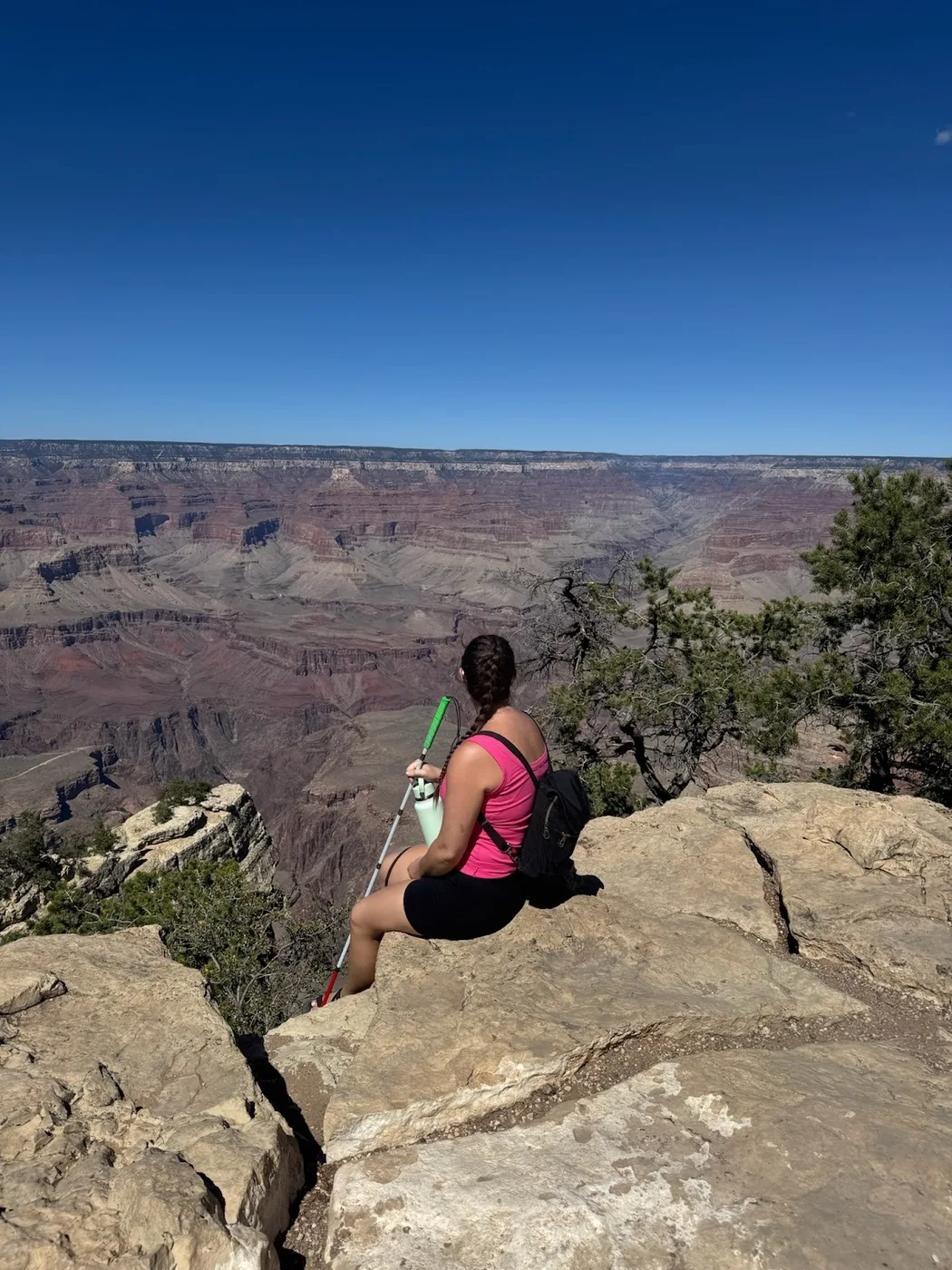 A photo of Monica sitting on a rock, holding her white cane, facing the Grand Canyon. Her back is to the camera, unaware that the photo is being taken.