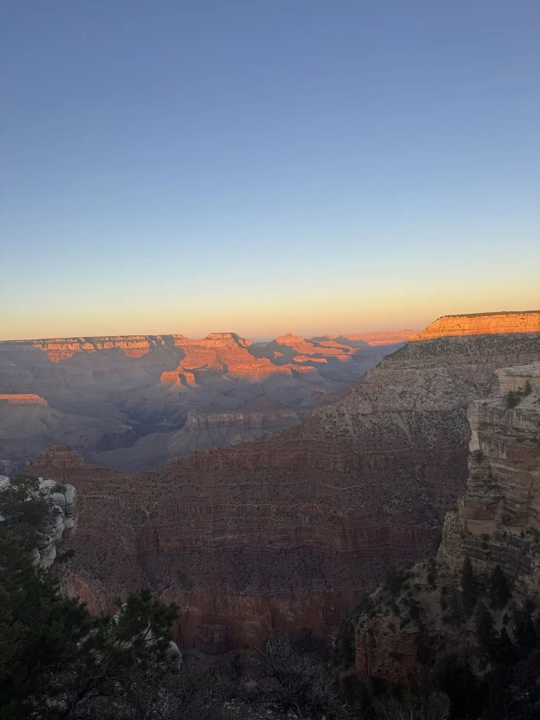 A photo of the Grand Canyon at sunset facing east. The sky is blue at the top with orange hues closest to the rocks. The top of the rocks are a vibrant red while the rest of the canyon is absorbed in shadows.