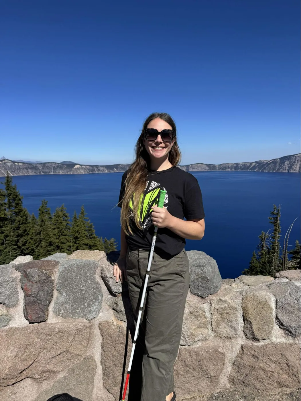 A photo of Monica standing with her white cane in front of a rock wall with Crater Lake behind her. The waters are a deep blue, which perfectly mirrors the clear, deep blue skies. She's wearing green hiking pants, an Awarewolf t-shirt, sunglasses, and hiking sandals.