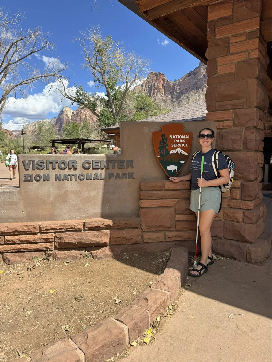 A photo of Monica standing next to the “Visitor Center of Zion” sign. She's holding her white cane and wearing an Usher Syndrome Awareness T-shirt.
