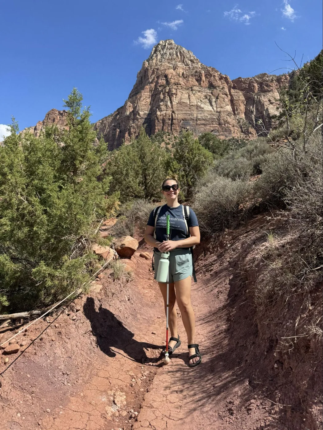 A photo of Monica standing, smiling, in front of a mountain at Zion. She's standing on a sandy path lined with rope, with shrubs lining the path on either side. She's holding her water bottle and white cane.