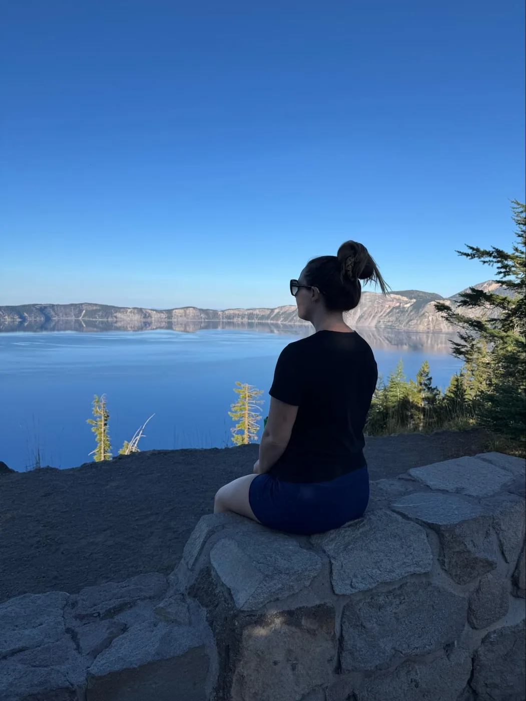 A photo of Monica sitting admiring Crater Lake. The sky is a deep blue, perfectly mirroring the clear blue waters of Crater Lake. She's facing away from the camera, wearing all black, sunglasses, and her hair is in a messy bun.