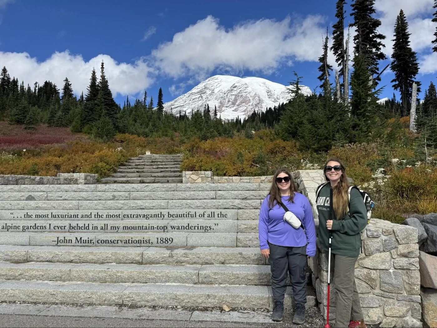 A photo of Monica and Erin standing on the stairs leading up to Skyline Loop with Mount Rainier in the background. The stairs have a quote from John Muir on them that reads: "... the most luxuriant and the most extravagantly beautiful of all the alpine gardens I ever beheld in all my mountain-top wanderings." —John Muir, conservationist, 1889.