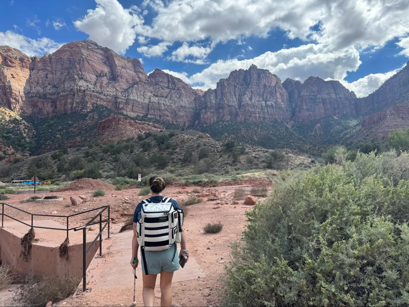 A landscape photo of the red rocks of Zion towering up with clouds in the sky. Monica’s back is to the camera, and she’s wearing her white FEELDOM backpack and leading with her white cane. She’s following a sandy path, and there are shrubs at the foot of the mountains.