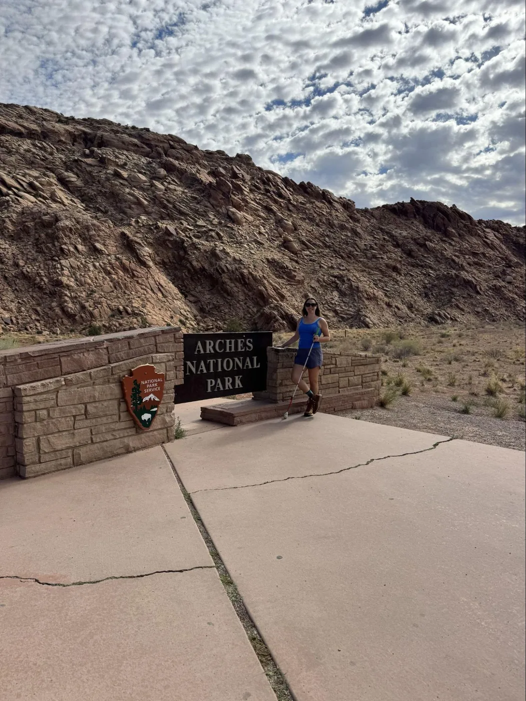 A photo of Monica, wearing a blue tank top and black shorts while holding a white cane, standing in front of the Arches National Park sign on a cloudy day.