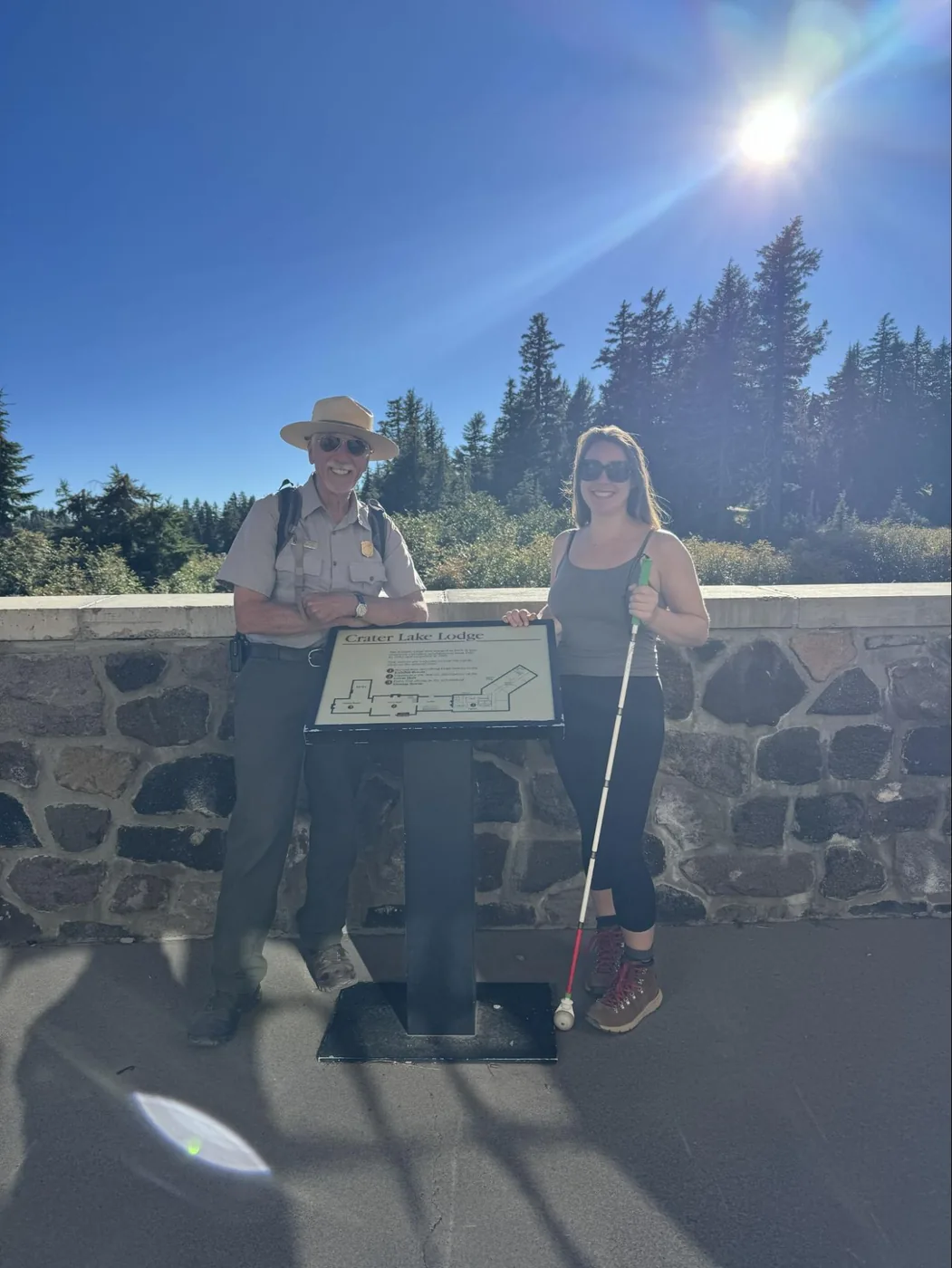 A photo of Ranger Bruce (L) and Monica (R) standing next to a placard that says Crater Lake Lodge on a sunny day with blue skies and green pine trees in the background. Ranger Bruce is an older white male with a mustache. He's wearing a wide-brimmed hat, ranger attire, and sunglasses. Monica is wearing sunglasses, a grey tank top, black leggings, and hiking boots. She's holding her white cane.