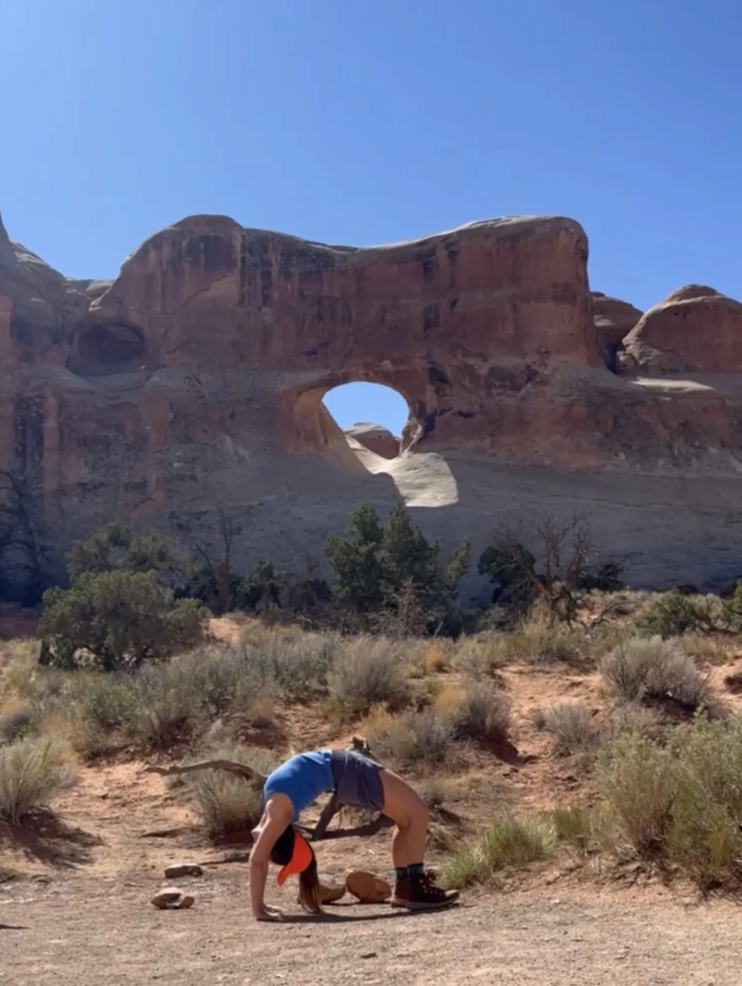 A screenshot from a video of Monica doing a bridge yoga pose in front of a tunneled, arched rock.