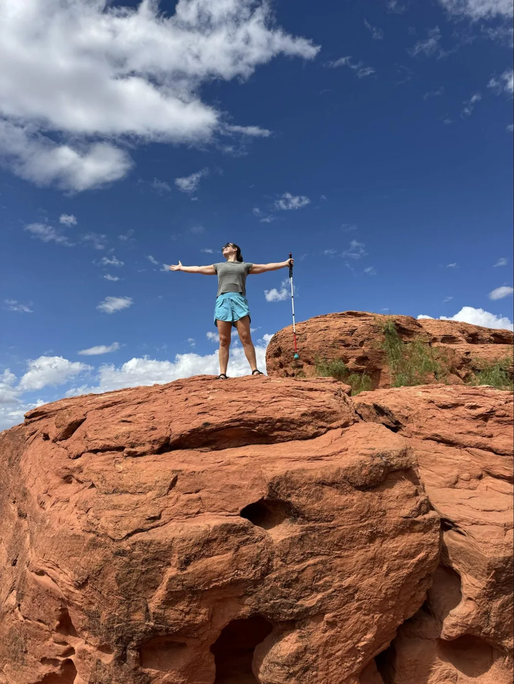 A photo of Monica standing on top of a red rock. The sky is blue with a few white clouds. She's standing tall with her arms out wide, looking out to the side. She's holding her white cane in one hand.