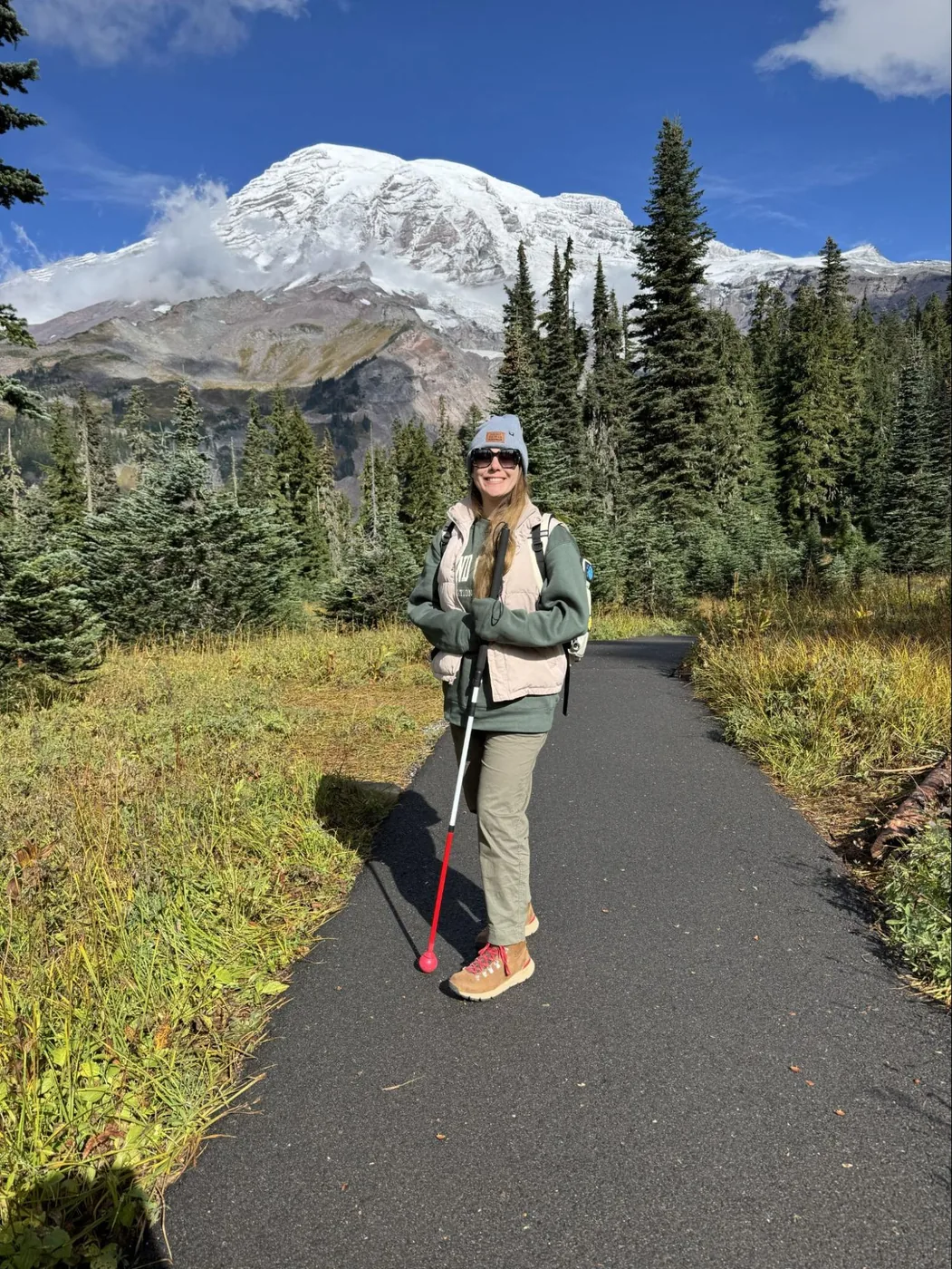 A photo of Monica standing with her all-terrain white cane on a paved trail with Mount Rainier behind her. It is a mostly clear blue sky with some small clouds forming around the glaciers on the mountain. She’s wearing a beanie, sunglasses, a pink puffer vest, a green sweatshirt, hiking pants, and hiking boots.