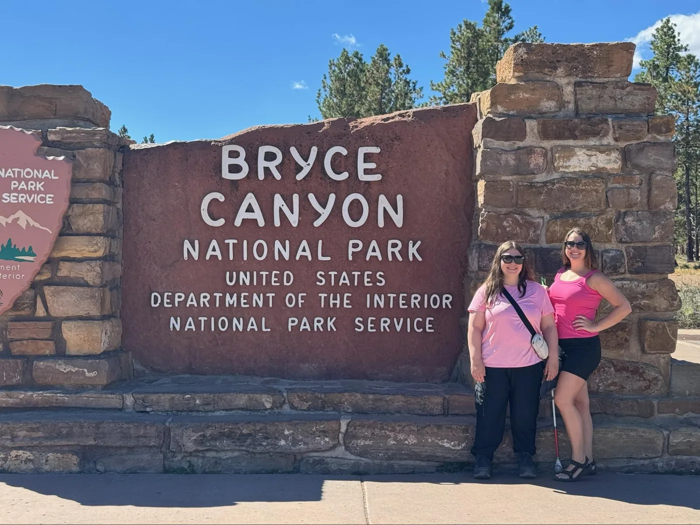 A photo of Erin and Monica standing next to the Bryce Canyon National Park sign. They're both wearing pink shirts and black bottoms.