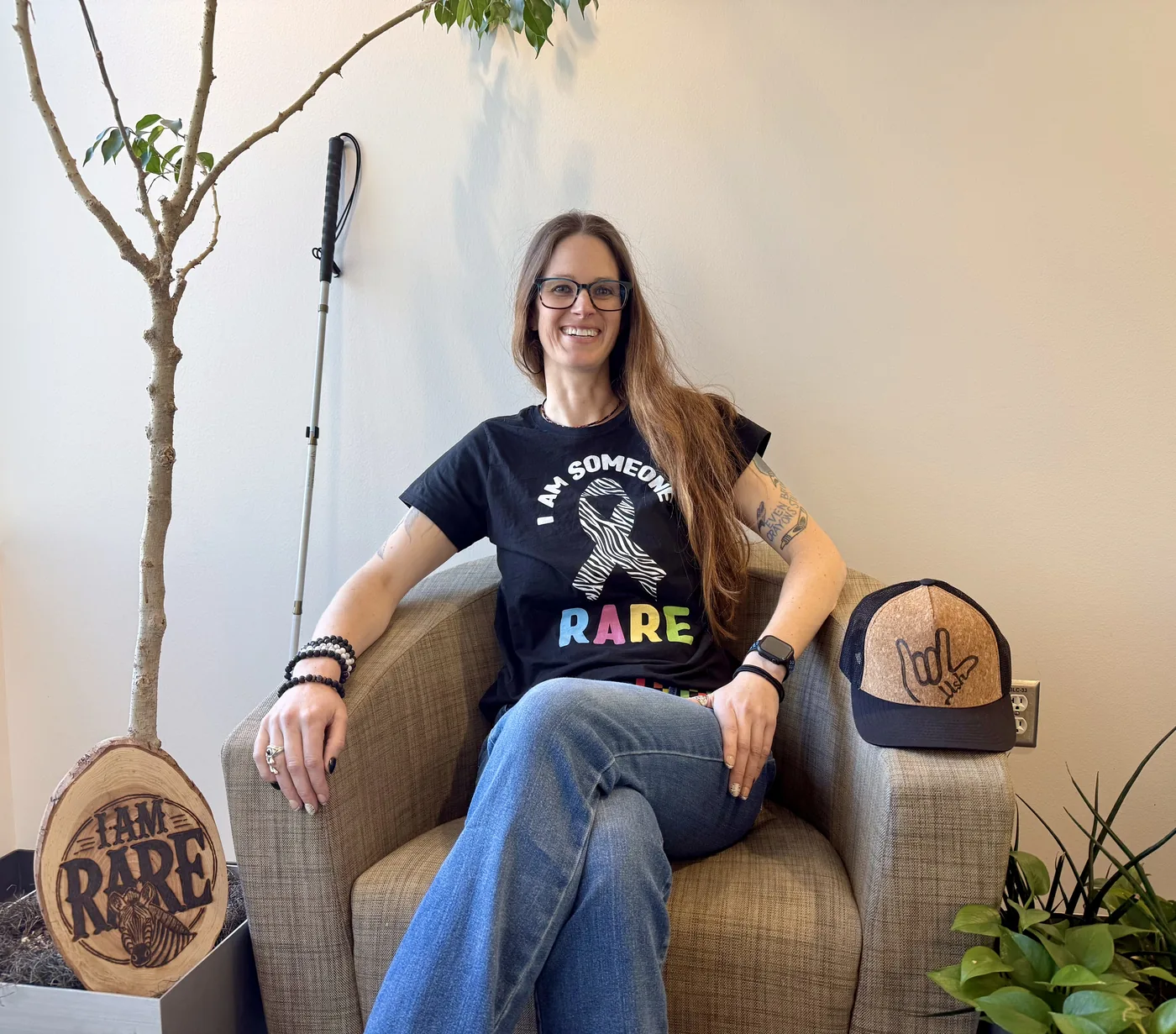 A woman with long brown hair and glasses sits smiling in a woven armchair, wearing a black “I AM SOMEONE RARE” t-shirt with a zebra-print awareness ribbon and blue jeans. She has tattoos on her left arm, several bracelets, rings, and a smartwatch. Beside her is a white cane leaning against the wall, a cork-and-black trucker cap with an “I love you” ASL hand sign on it alongside a signature, and a wood slice sign that is wood burned with “I AM RARE” and a zebra illustration, near a small potted tree. She’s in a room with light colored walls and green potted plants.