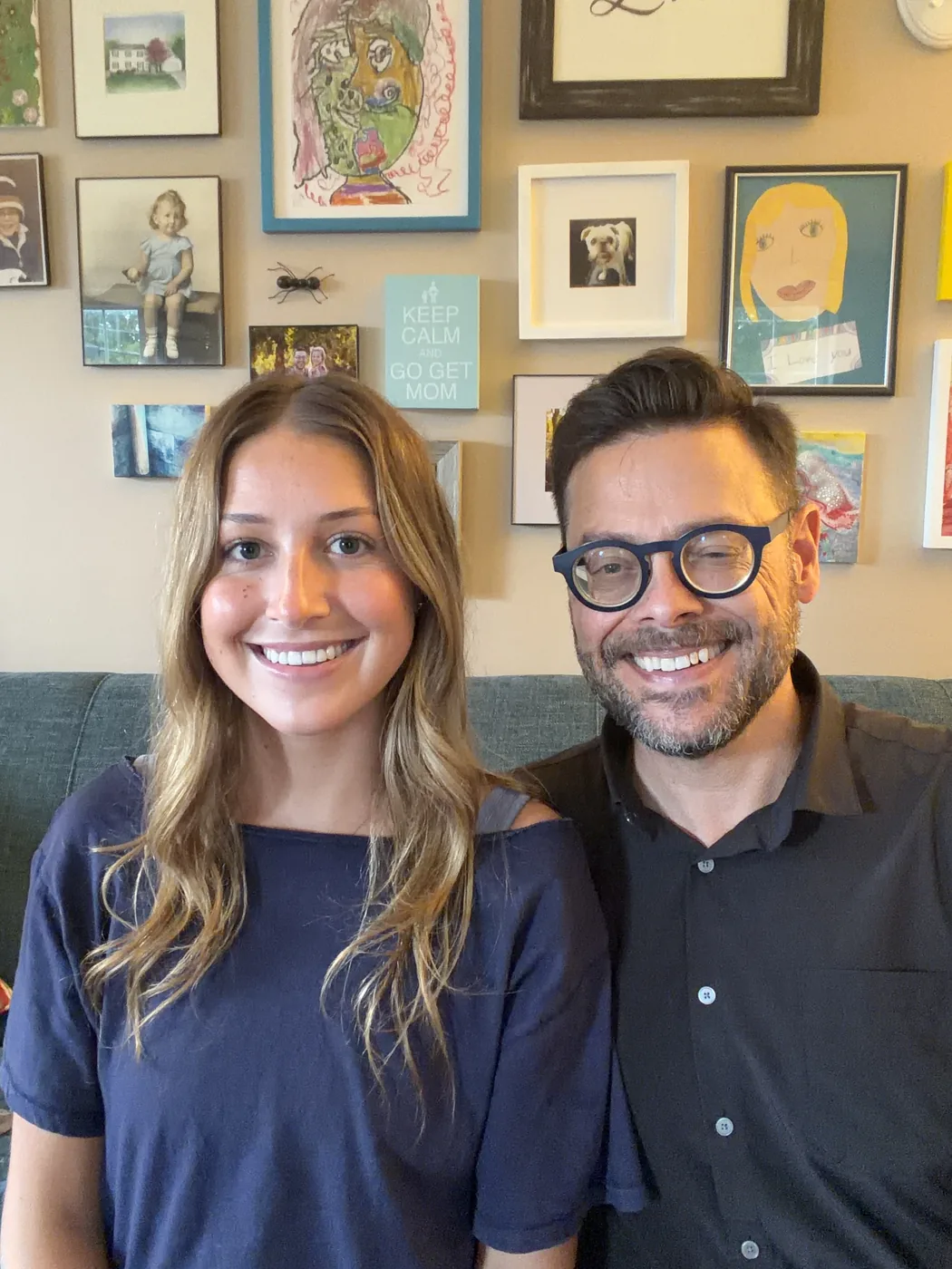 Father and daughter, Josh and Eileen Loebner excitedly smiling, sitting together on a couch with whimsical family artwork framed on the wall behind them.