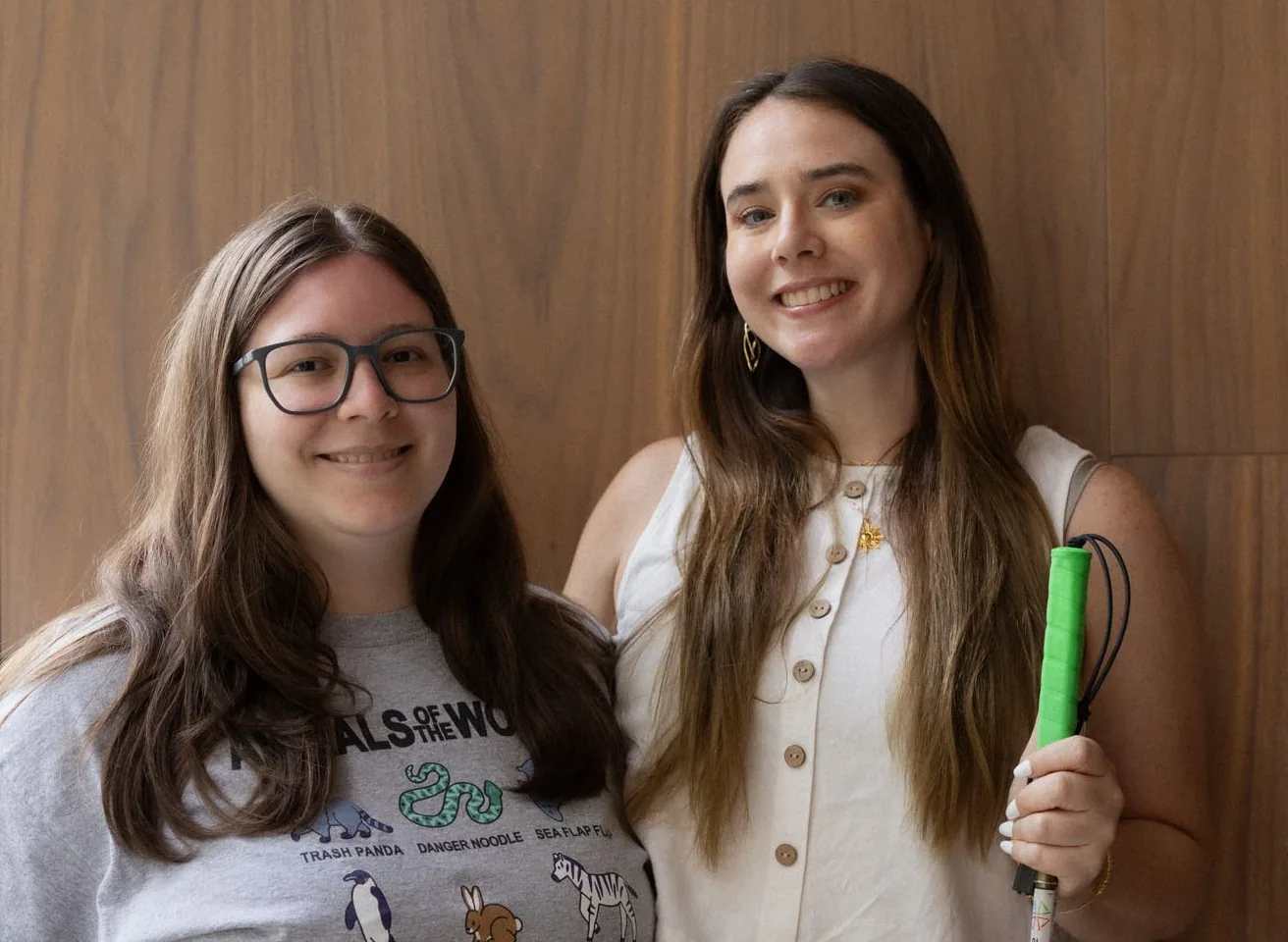 Monica and Erin Pruitt standing together against a wood-panelled wall, smiling at the camera. One of them holds a white cane with a bright green grip.