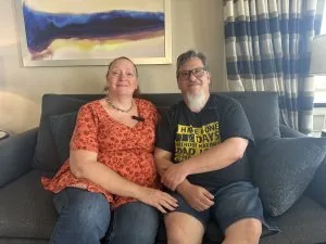 Ted and Carrie Tahquechi sitting side by side on a grey sofa beneath a framed painting.