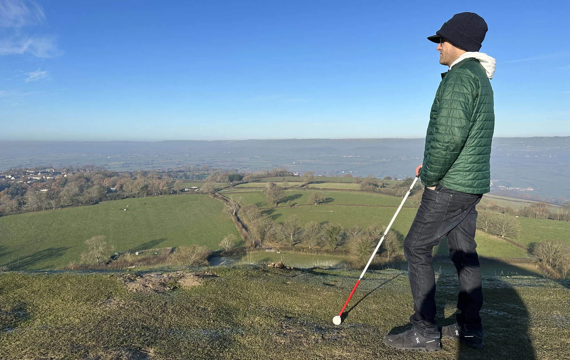 A traveller in a dark green jacket stands on a grass ridge, white cane angled down into the turf, facing a wide valley of hedgerows, bare trees and pale estuary light.