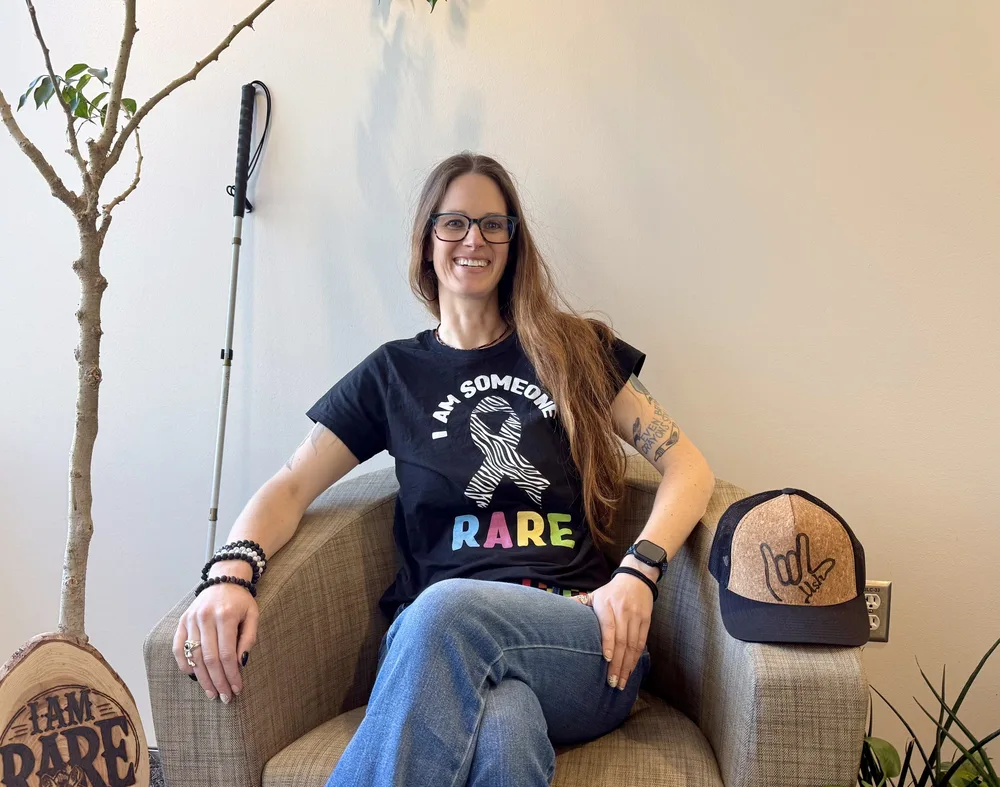 A woman seated in an armchair beside a potted tree, wearing a shirt reading I am someone RARE.