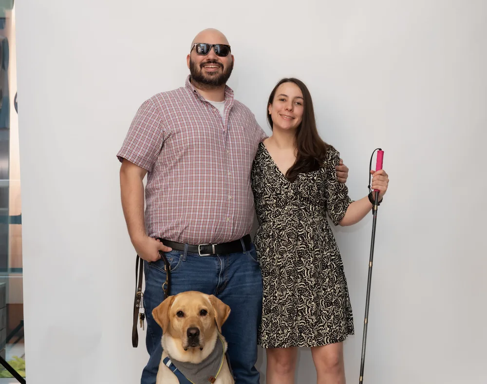 Emeline Lakrout and Daniel Gutz standing with their yellow guide dog, one holding a white cane.