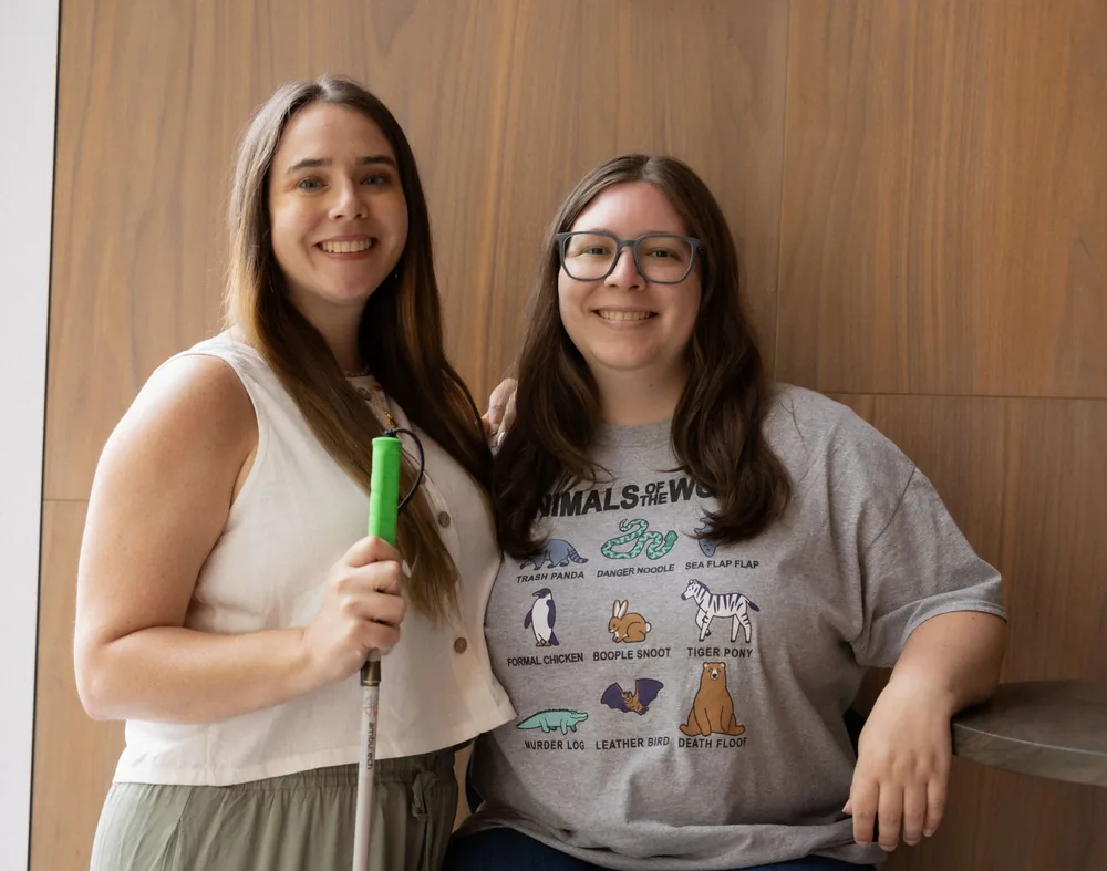 Erin and Monica Pruitt sitting together against a wood-panelled wall, one holding a white cane with a green grip.