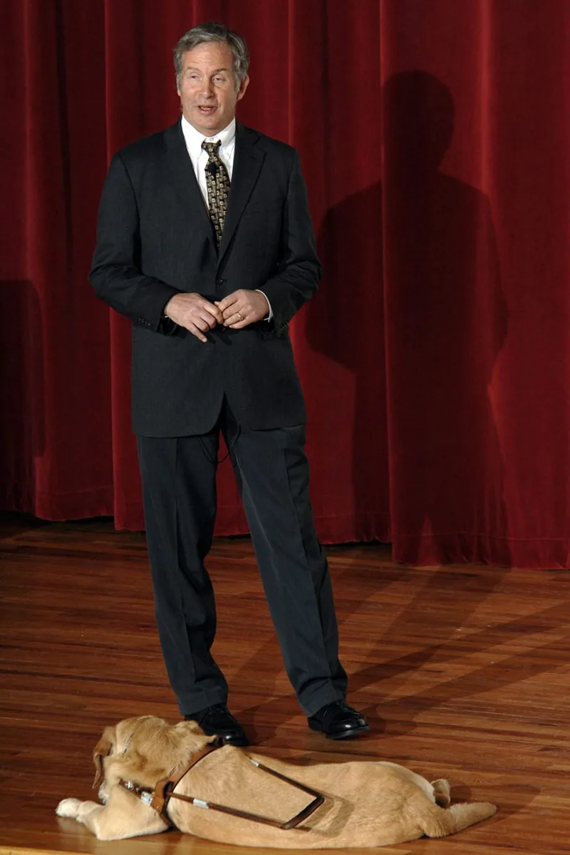 A speaker in a dark suit on a stage with a red curtain, his guide dog lying at his feet.