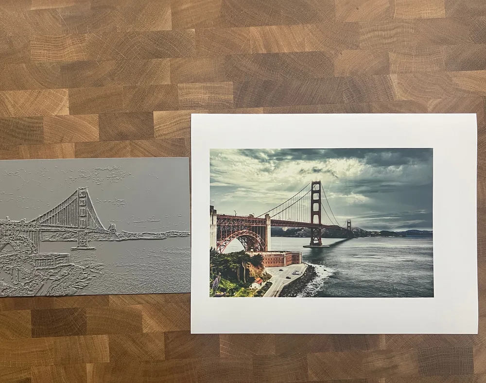 A raised-line tactile print of the Golden Gate Bridge lying on a wooden table beside the photograph it was made from.
